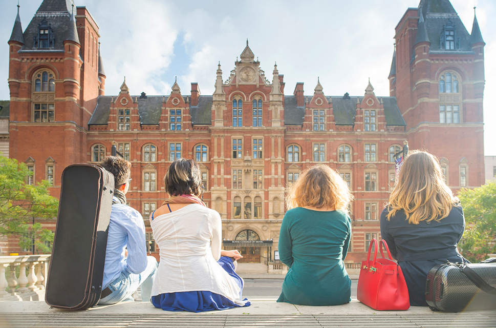 A group of students, with bags and music cases next to them, sitting on some steps, facing an old red-brick building, the Royal College of Music main building.